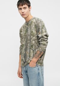 Μακρυμάνικο πουκάμισο με καμουφλάζ σε πράσινο και καφέ χρώμα, υφασμάτινη υφή που μοιάζει με φλοιό δέντρου, σε συνδυασμό με ανοιχτό μπλε τζιν.