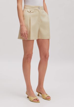 Vrouw draagt beige shorts met hoge taille en knoopsluiting aan de zijzak, gecombineerd met beige open-toe haksandalen en een wit mouwloos topje.