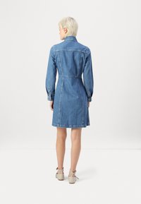 Jeans haljina u plavoj boji, s dugim rukavima, uskim gornjim dijelom i raširenim suknjom. Detalji uključuju ovratnik i manšete s dugmadi.