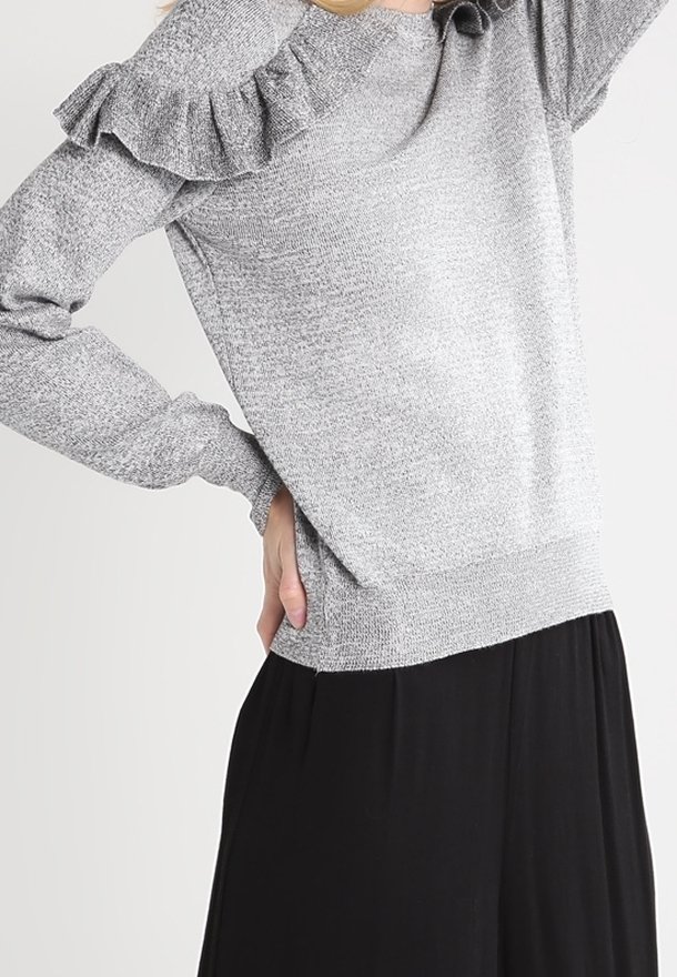 Femme portant un pull gris en tricot avec un détail de volants sur les épaules et un pantalon noir à jambes larges, posant avec une main sur la hanche et l'autre levée.