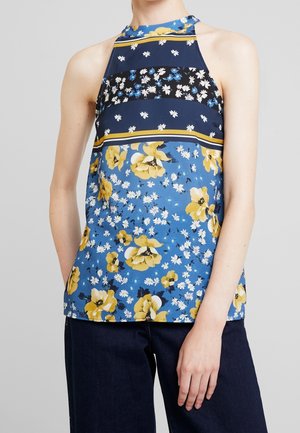 Mouwloos haltertop met blauwe, zwarte en gele bloemenstrepen, gedragen met donkere broek, tegen een effen lichtgrijze achtergrond.