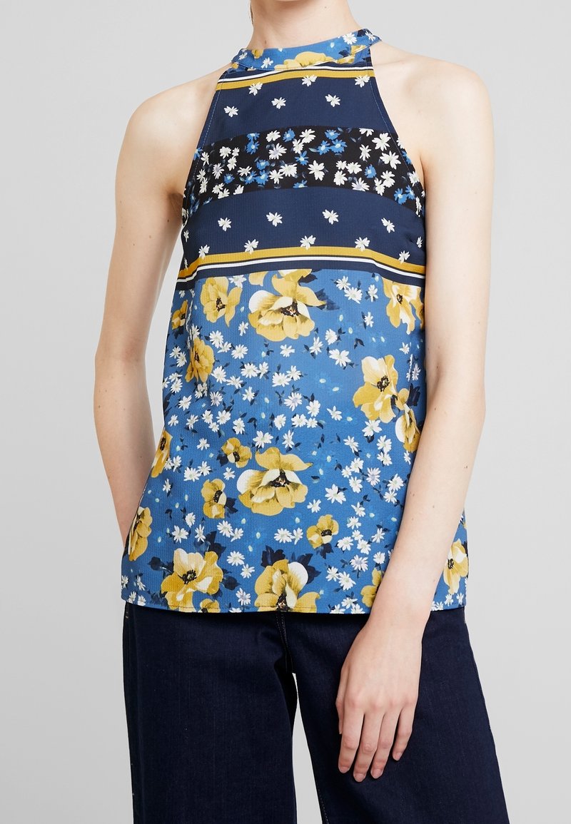 Haut sans manches à col licou avec des rayures florales bleu, noir et jaune, porté avec un pantalon foncé, sur un fond gris clair uni.