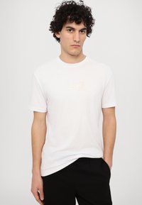 Hvid bomulds-T-shirt med korte ærmer, prydet med et struktureret gyldent "EA7" logo på brystet. Modellen bærer sorte bukser.