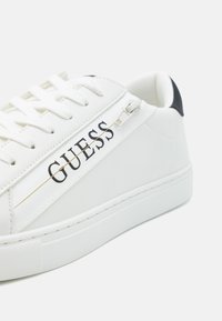 Fehér sneaker sima szintetikus bőr felsőrésszel, fekete kiegészítővel a sarkán, fűzőzáró rendszerrel és fényes logóval az oldalon. Texturált talp.