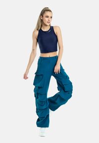 Pantaloni cargo verde acqua con molte tasche grandi e un design svasato, abbinati a un top corto blu navy, indossati con sneakers bianche.