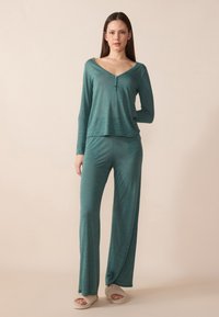 Teal loungewear set with long sleeves, V-neck, and 3-button detail; smooth texture; relaxed fit; paired with light-colored slippers.