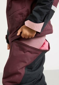 Bordeaux en zwart winterpak met een ritssluitingzak die een roze voering onthult, voorzien van contrasterende mouwaccenten en een gestructureerde stof.