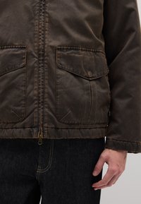 Braune Reißverschlussjacke mit zwei aufgesetzten Fronttaschen, aus strukturiertem Material, mit sichtbaren Nähten und einem weichen, leicht getragenen Aussehen.