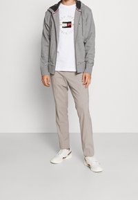Grau zip-up Hoodie mit schwarzer Innenausstattung, weißes T-Shirt mit Logo, beigefarbene Hose und weiße Sneakers mit roten Akzenten. Schlichter, lässiger Look.
