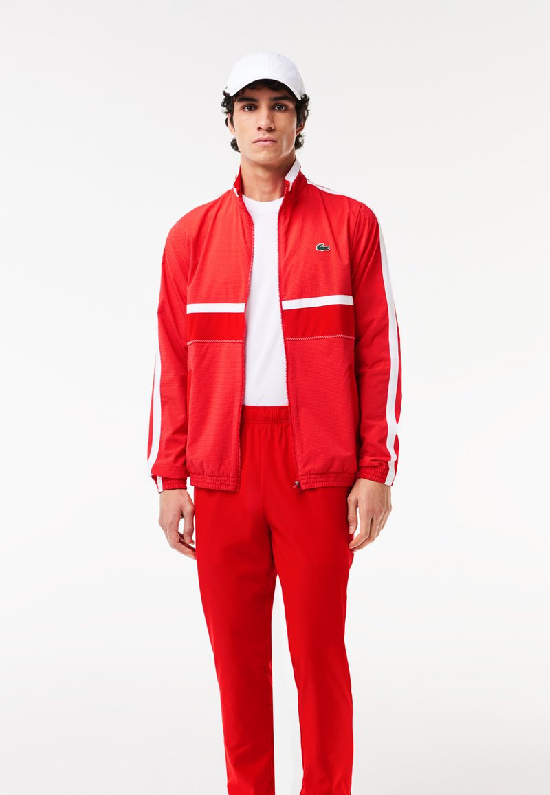 Veste de sport rouge avec des rayures blanches, fermeture zip, et col montant, accompagnée d'un pantalon rouge assorti. Une casquette blanche complète la tenue.