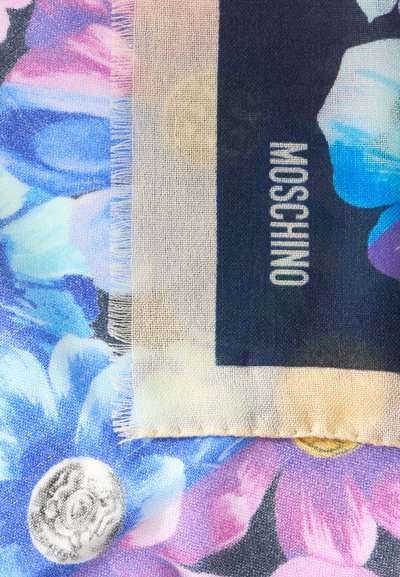 Écharpe avec une bordure beige et un centre foncé avec le texte "MOSCHINO", posée sur un tissu à fleurs bleues, roses et violettes.