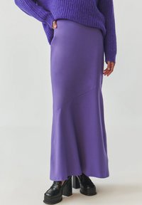 Violetti maxi-hame, joka on valmistettu sileästä ja joustavasta kankaasta. Tässä on kaareva sauma sekä kainalosilhuetti, joka on yhdistetty mustiin korkokenkiin.