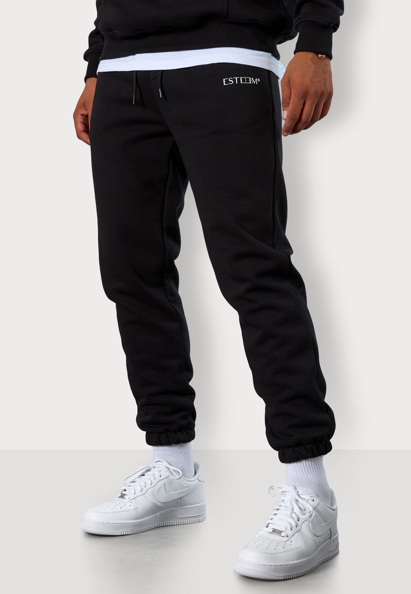 Schwarze Sweatpants mit elastischem Bund und Bündchen, versehen mit einem dezenten "ESTEEM"-Logo. Kombiniert mit weißen Sneakers und weißen Socken.