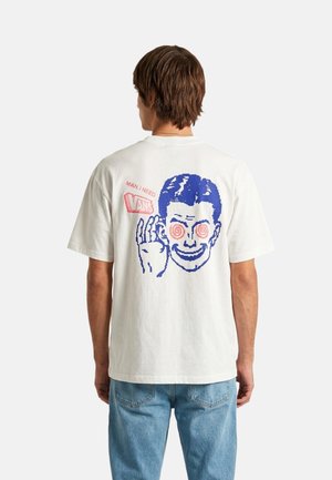 Mann trägt weißes T-Shirt mit großem blauem Cartoon-Gesicht mit Spiralaugen und Text „MAN, I NEED VANS“ auf dem Rücken, kombiniert mit hellblauen Jeans.