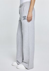 Lichtgrijze wijde sweatpants van zachte stof, met een elastische tailleband en een logo-opdruk op de linkerdij. Gecombineerd met witte schoenen.