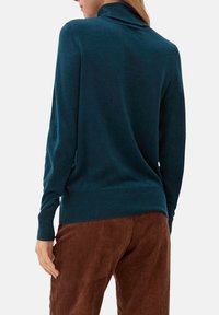 Pull long turquoise à manches longues avec un col et des poignets côtelés ; texture douce et silhouette ajustée, associé à un pantalon en velours côtelé marron.