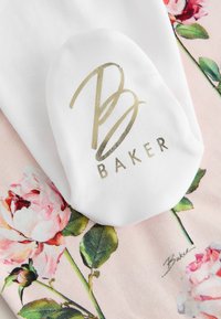 Tessuto bianco con logo dorato "B Baker" su tessuto rosa con motivi floreali di rose e foglie verdi.