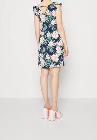 Femme portant une robe florale bleu marine avec des fleurs roses, des manches volantes, une découpe dans le dos, et des sandales à plateforme roses à talons hauts, se tenant sur un sol blanc.