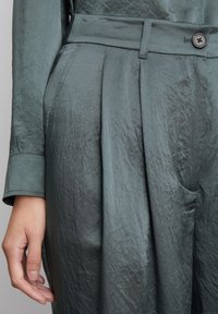 Pantalons en tissu vert, froissés, avec un design plissé. Comprend un bouton visible et une ceinture, mettant en valeur une texture lisse et une coupe ajustée.