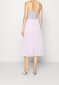 Mujer de pie de espaldas, vistiendo un vestido midi sin mangas color lavanda con un corsé adornado y sandalias blancas de tacón.