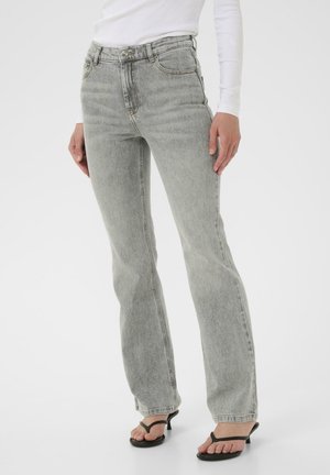 Kvinde iført lysegrå jeans med lige ben, hvid ribbet langærmet top og sorte sandaler med hæl, stående mod en hvid baggrund.