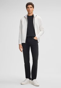 Weißer Zip-Hoodie mit grauem Kapuze, schwarzes T-Shirt, dunkle Jeans und weiße Sneakers. Hat eine körpernahe Passform und leichten Stoff.