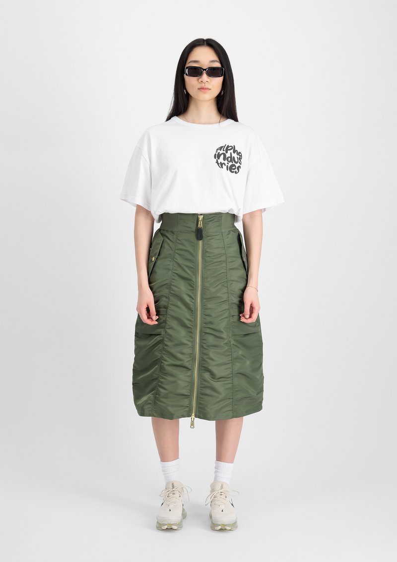T-shirt bianca oversize con logo nero, abbinata a una gonna midi trapuntata verde con zip frontale e tasche laterali, indossata con sneakers.
