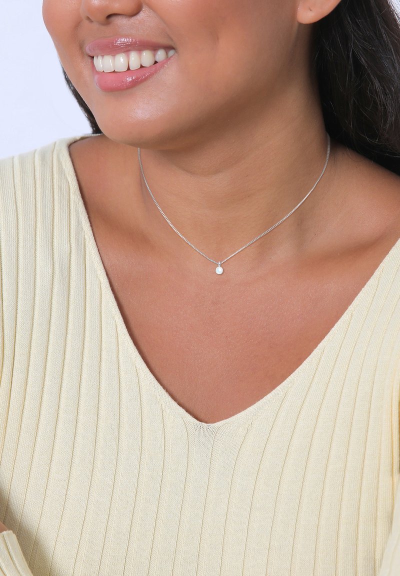 Collana d'argento con un piccolo pendente rotondo, indossata su un collo nudo. La parte superiore è di un giallo chiaro, con una texture a righe verticali.
