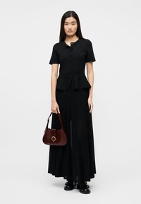 Γυναίκα με μακριά σκούρα μαλλιά, που φοράει μαύρο κοντομάνικο τοπ με κουμπιά και μακριά φούστα, κρατώντας μια σκούρα κόκκινη τσάντα, στέκεται σε λευκό φόντο.