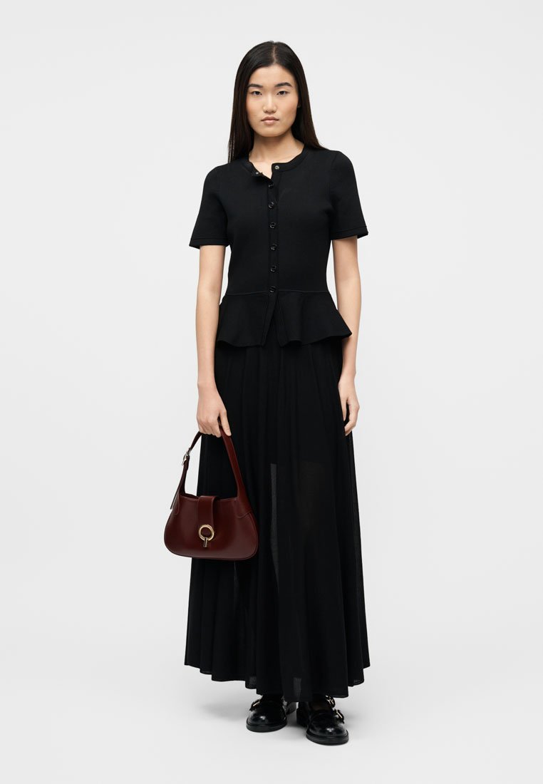 Femme aux longs cheveux foncés portant un haut noir à manches courtes boutonné et une longue jupe, tenant un sac à main rouge foncé, debout sur un fond blanc.
