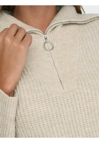 Pull en maille beige clair avec un col montant, présentant une fermeture à demi-glissière et un tirette de zip circulaire en argent, texturé avec un motif côtelé.