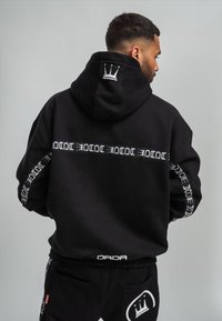 Schwarzer Kapuzenpullover mit großem Rückenlogo und weißer Beschriftung auf den Ärmeln. Verfügt über eine Kapuze mit Kordelzug und eine Kängurutasche. Weiche Stoffstruktur.