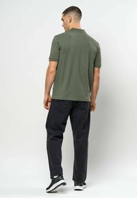 Mann som står med ryggen til, iført en olivengrønn poloshirt, svarte jeans og svarte og hvite joggesko mot en ensfarget hvit bakgrunn.