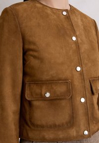 Braune Wildlederjacke mit rundem Ausschnitt, silbernen Druckknöpfen und zwei vorderen Taschen mit Klappen. Glatte Textur und taillierte Passform.