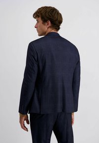 Mann steht nach links gewandt, trägt einen dunkelblauen karierten Anzug und ein senfgelbes Langarmhemd unter dem Jackett.