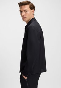 Black lightweight jacket with a zip closure, long sleeves, a chest pocket, and buttoned cuffs, featuring a smooth texture and tailored fit.