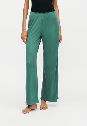 Femme portant un pantalon ample côtelé couleur sarcelle, debout pieds nus sur un fond blanc uni.