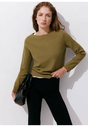 Mujer con cabello castaño claro que lleva un suéter verde oliva y pantalones negros, sostiene un bolso negro frente a una pared blanca.