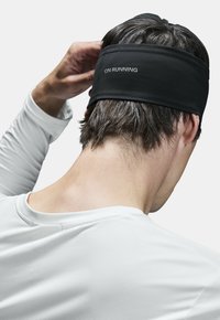 Diadema negra para correr con el logo "ON RUNNING". Tejido elástico, diseño elegante. Llevada en la cabeza, con el cabello visible en la parte de atrás.