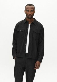 Black button-up jacket with two chest pockets, made from textured fabric. Worn over a white shirt, displaying a simple and modern design.