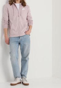 Sudadera con cremallera de color rosa claro con un logo, llevada sobre una camiseta blanca, combinada con jeans de color azul claro y zapatillas blancas con detalles en marrón.