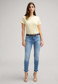Hellgelbes kurzärmeliges T-Shirt kombiniert mit hellblauen Skinny-Jeans, die einen ausgefransten Saum und einen schwarzen Gürtel mit einer dekorativen Schnalle aufweisen.