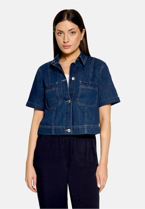 Femme portant une veste en jean bleu foncé à manches courtes avec des boutons sur le devant et deux poches poitrine, associée à un pantalon noir.