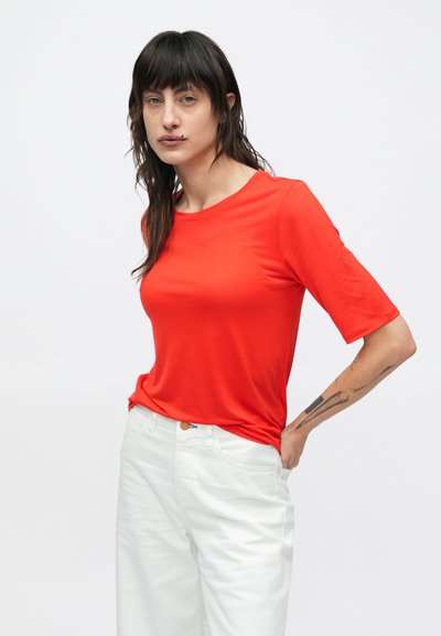 Piros, rövid ujjú póló puha anyagból, klasszikus kerek nyakkivágással és enyhén testhezsimuló fazonnal, fehér nadrággal párosítva.