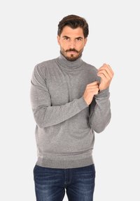 Maglione grigio a collo alto con polsini e scollo a costine, realizzato in materiale morbido, indossato con jeans blu, modello mentre sistema le maniche.