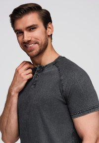 Šedé tričko s krátkým rukávem typu henley s knoflíkovou légou, kulatým výstřihem a jemnou texturou. Model, který má na sobě tričko, se lehce dotýká límečku.