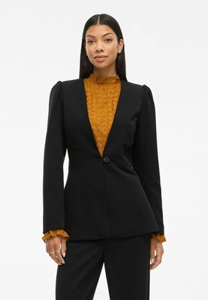 Blazer nero con scollo a V profondo, chiusura con bottone e spalle strutturate. Indossato sopra una blusa gialla a trama con colletto e polsini arricciati.