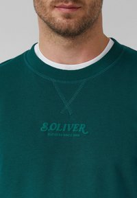 Felpa verde scuro con collo a giro, caratterizzata da cuciture testurizzate e un logo S.Oliver embossato di un verde più chiaro.