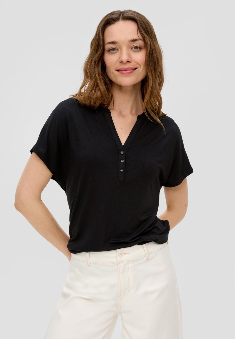 Zwarte blouse met korte mouwen en een V-hals, voorzien van een vierknopenpas. Gemaakt van zachte stof met een ontspannen pasvorm. Gecombineerd met lichte broek.