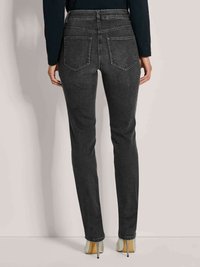 Vrouw in strakke zwarte jeans en zilveren hoge hakken, van achteren gezien tegen een effen lichte achtergrond.